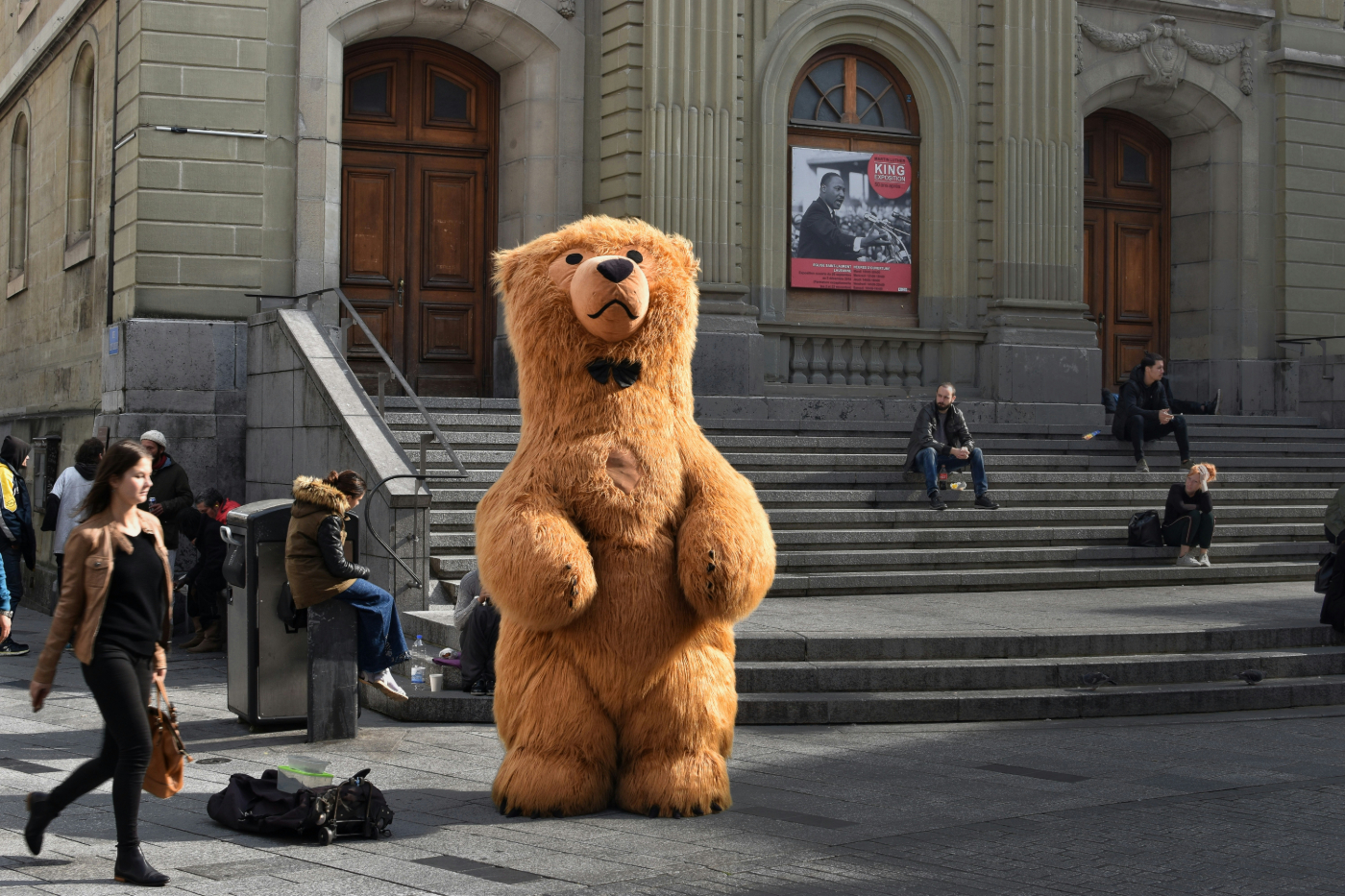 Someone dressed as a giant brown bear with a bow tie standing in front of a stone church
