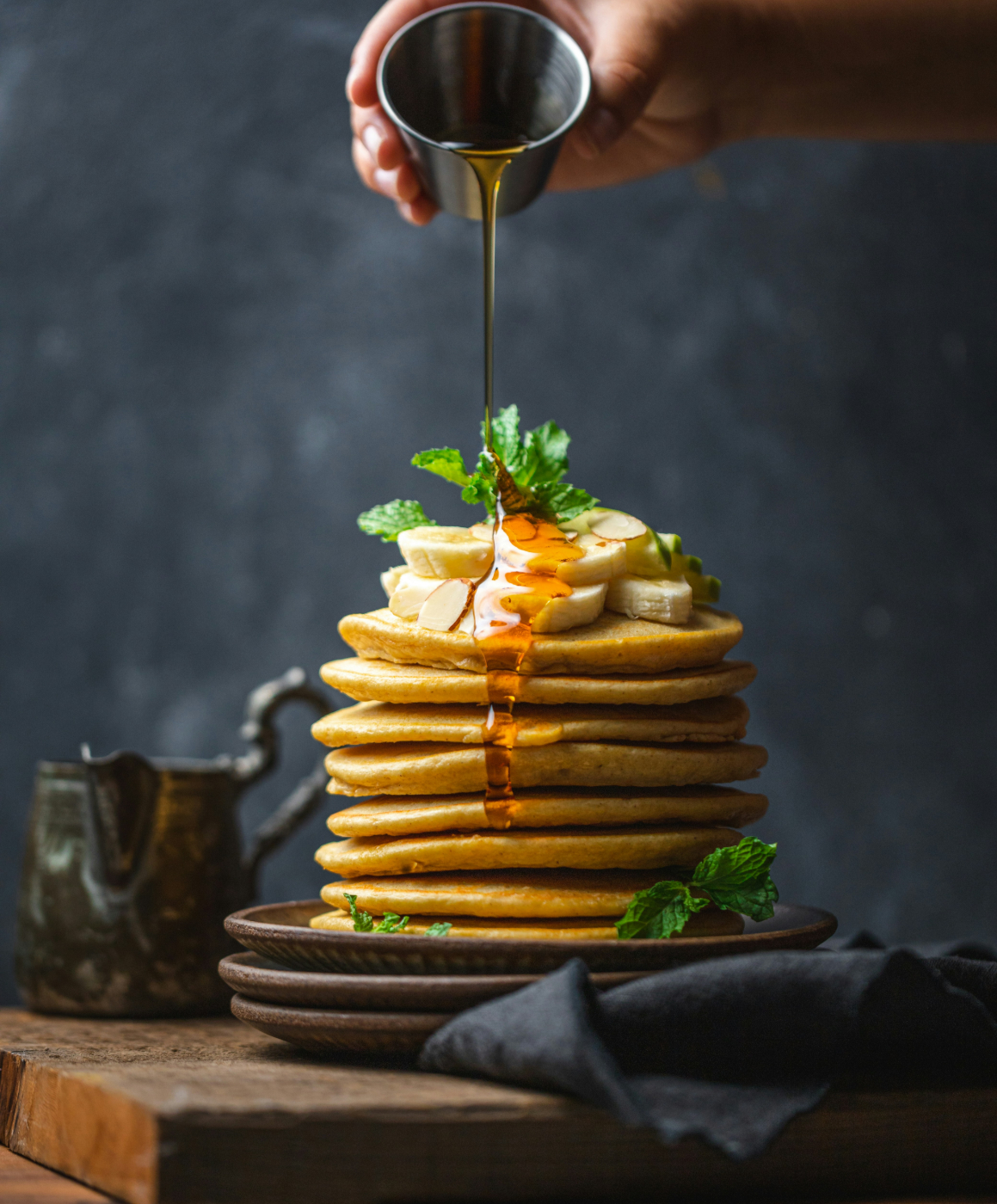 A stack of pancakes topped with sliced bananas and mint having syrup poured over them